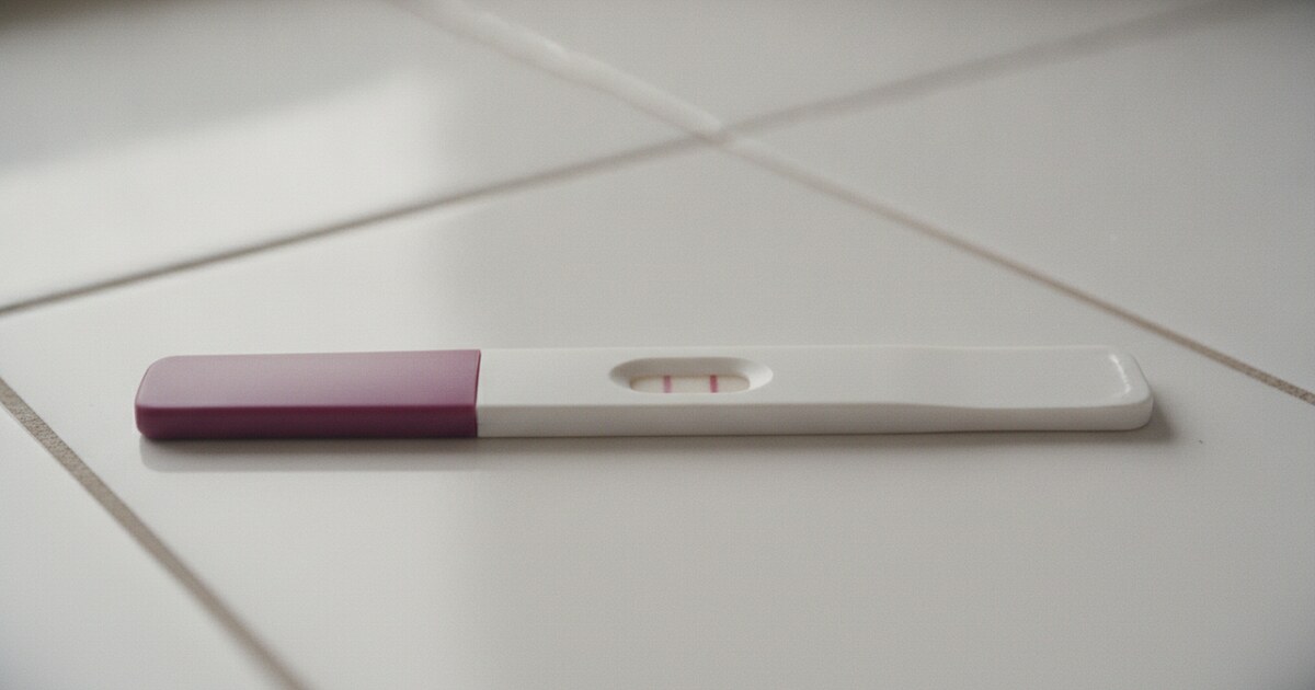 Home pregnancy test strip on a white tile surface in natural morning light, faint second line just visible in the test window