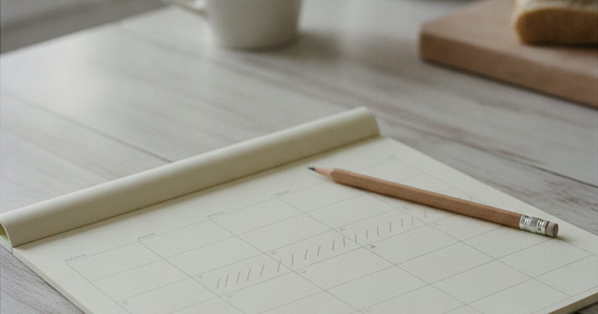 Paper wall calendar with small pencil tick marks counting off days, a pencil and a mug beside it on a kitchen table in soft daylight